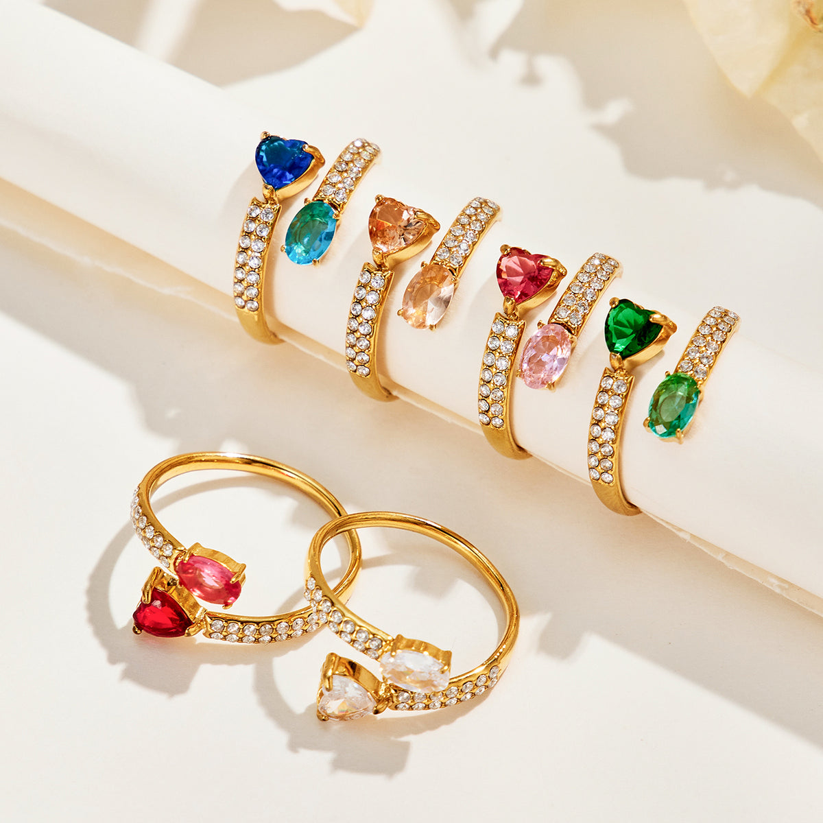 Gold-tone stainless steel heart and oval rings in multiple stone colors displayed along a cream ledge and on the surface below