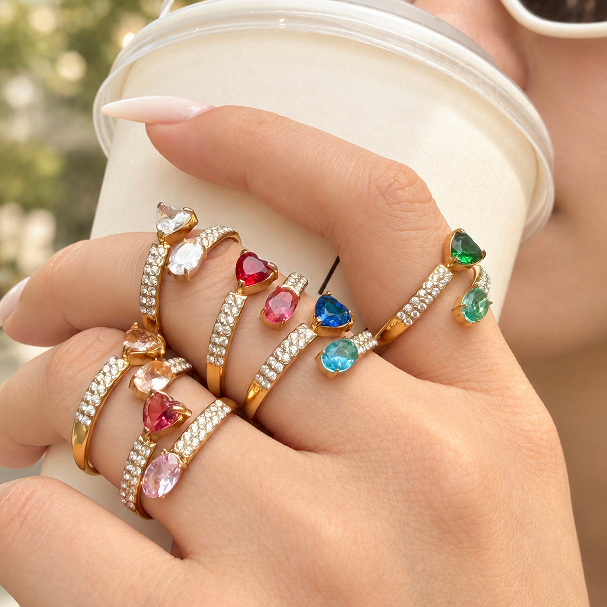 Gold-tone stainless steel heart and oval stone rings in multiple colors worn on a hand holding a white cup