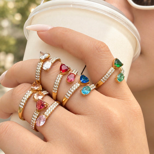 Gold-tone stainless steel heart and oval stone rings in multiple colors worn on a hand holding a white cup