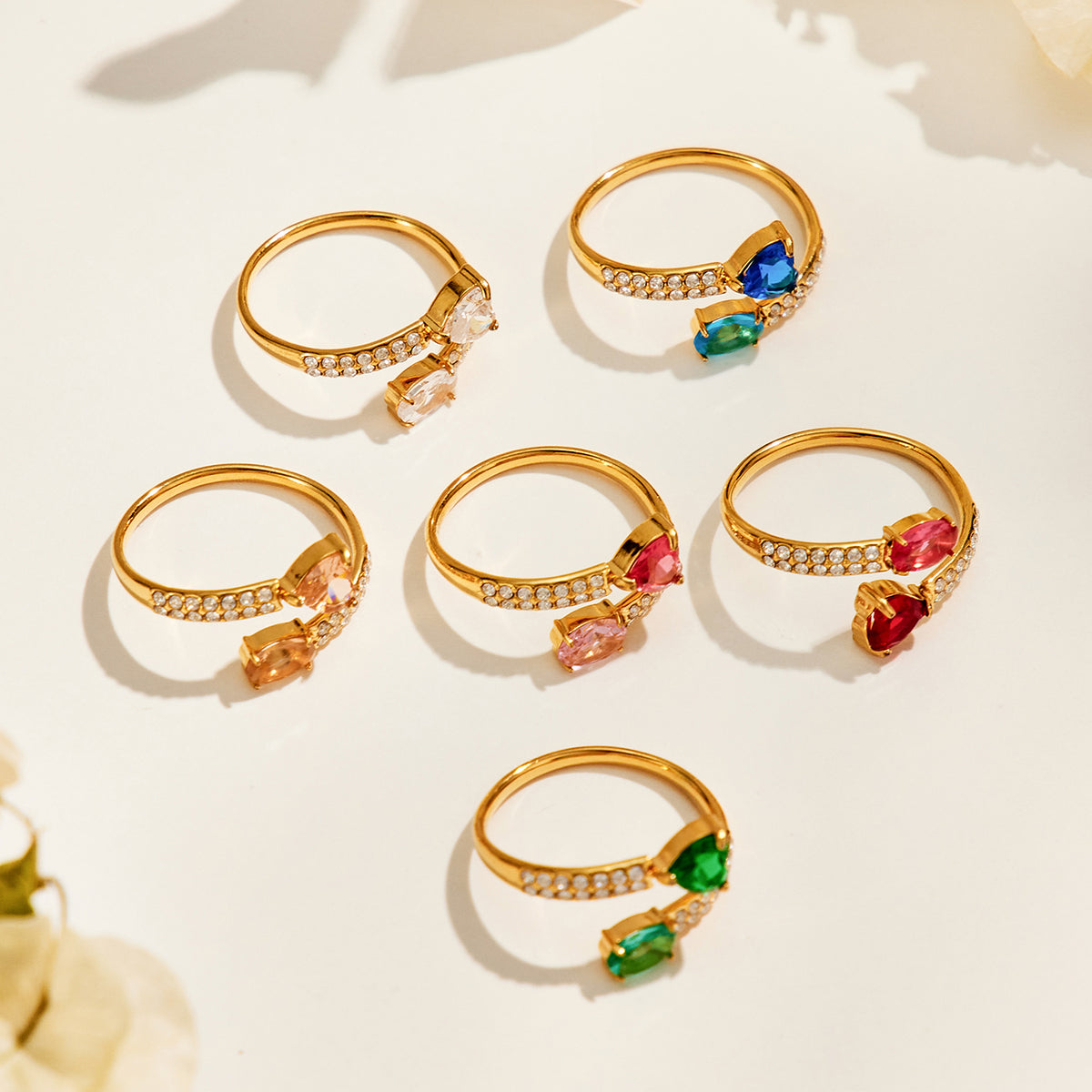 Six gold-tone stainless steel heart and oval rings arranged on a cream surface, showing their bands and colored settings from side and back angles