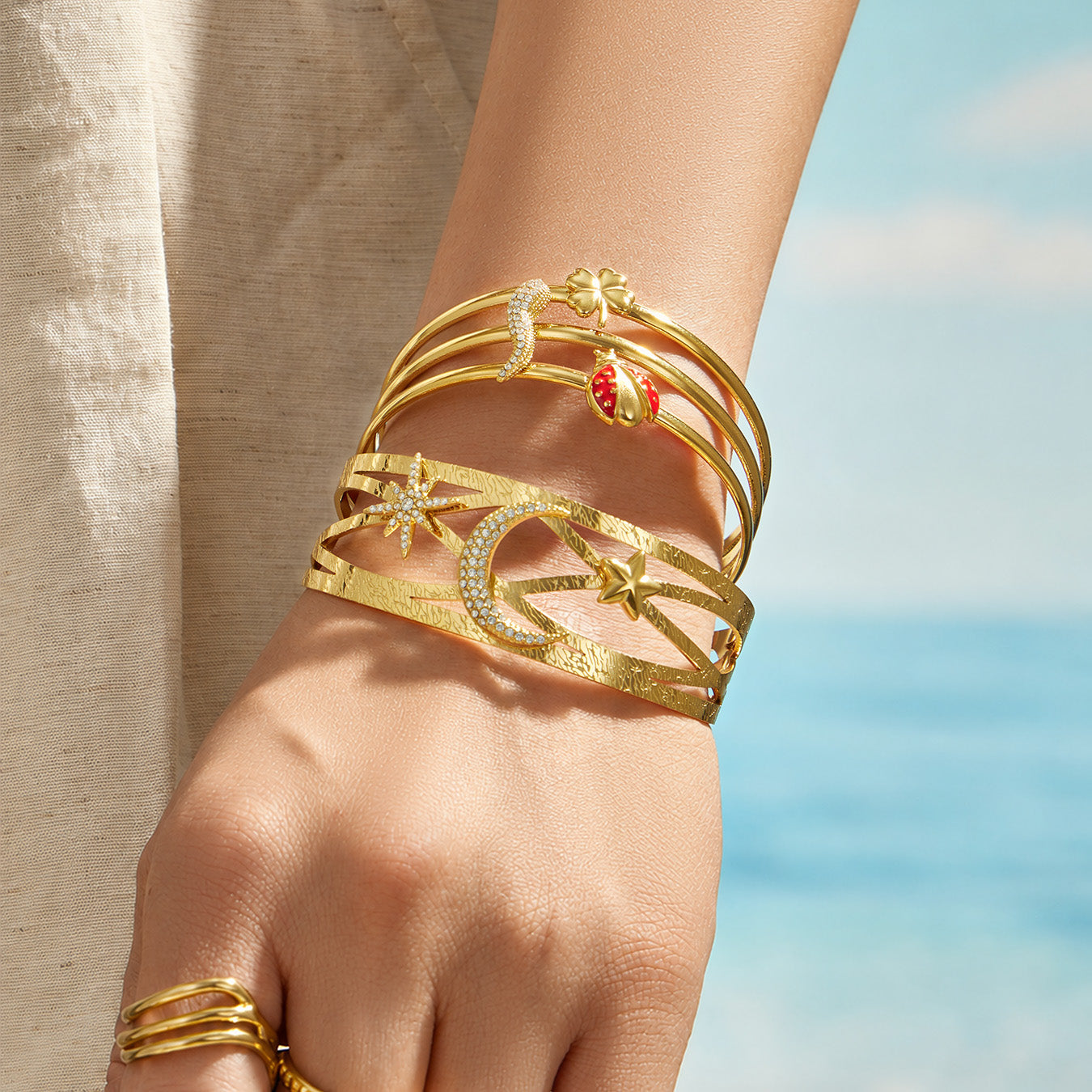Gold celestial and chili pepper cuff bracelets stacked on a model wrist by the sea