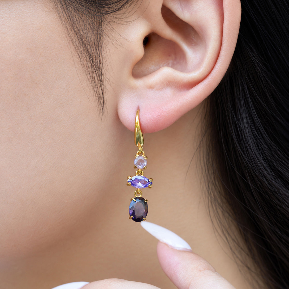 Close-up of a gold fishhook drop earring with purple and lavender stones, shown on a model's ear with a fingertip nearby