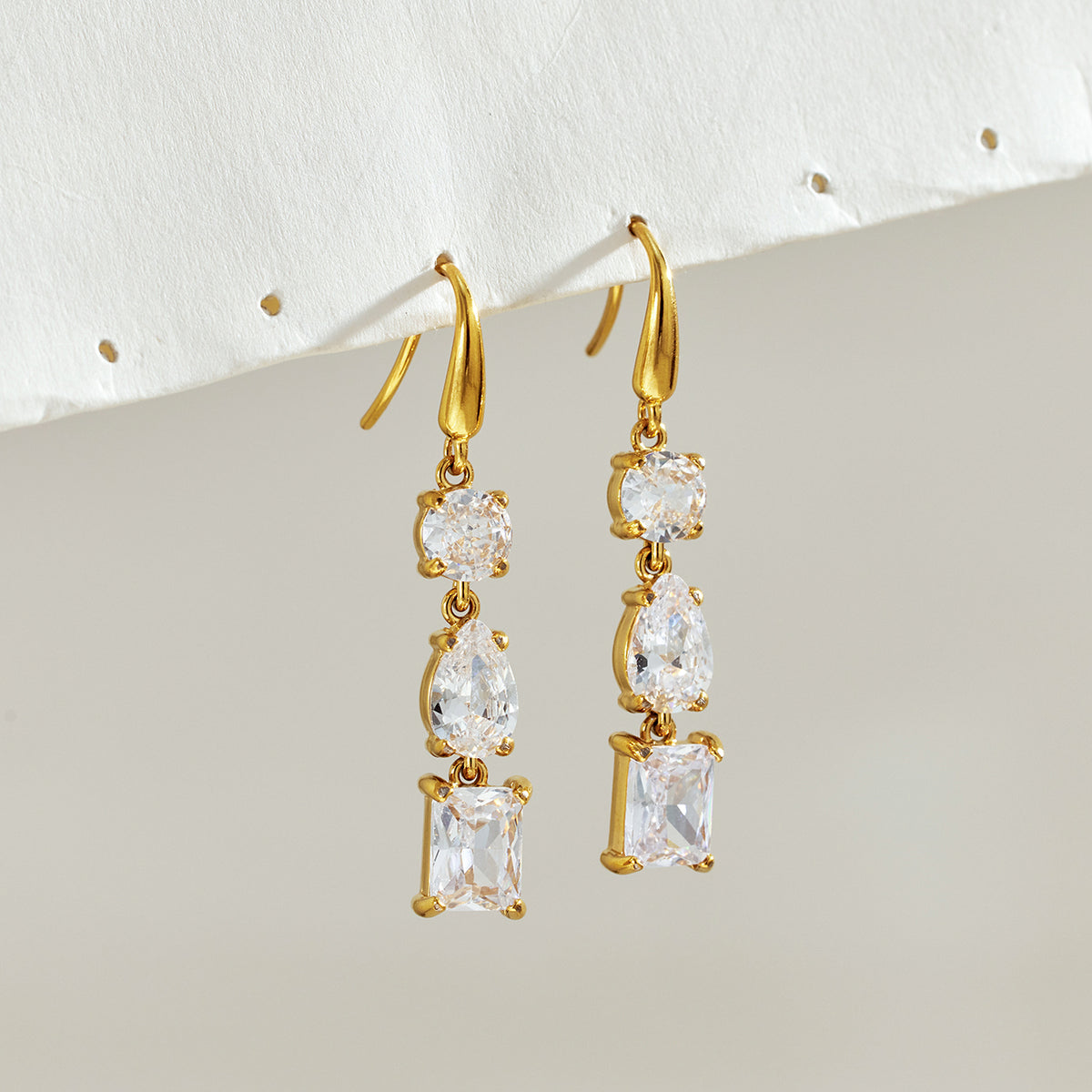 Pair of gold fishhook earrings with clear round, pear-shaped and rectangular stones hanging against a light background