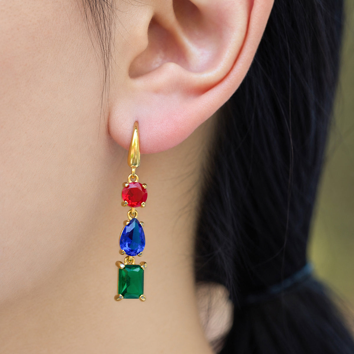 Model wearing a gold geometric dangle earring with red round, blue pear-shaped and green rectangular stones