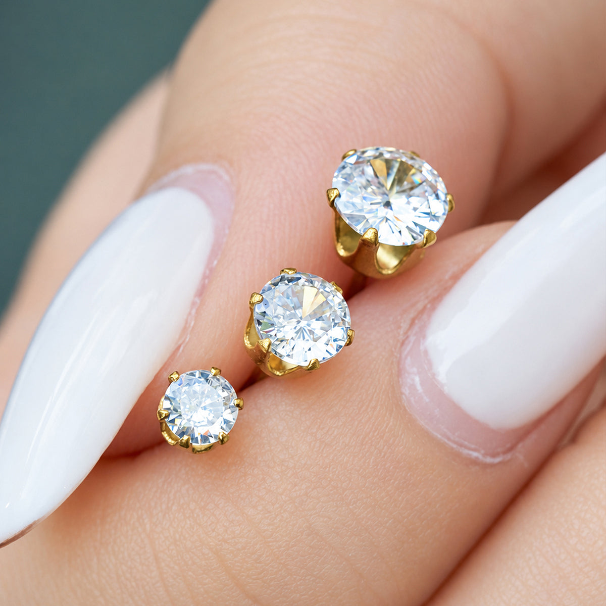Three gold-tone round solitaire stud earrings displayed between fingers to compare their stone sizes