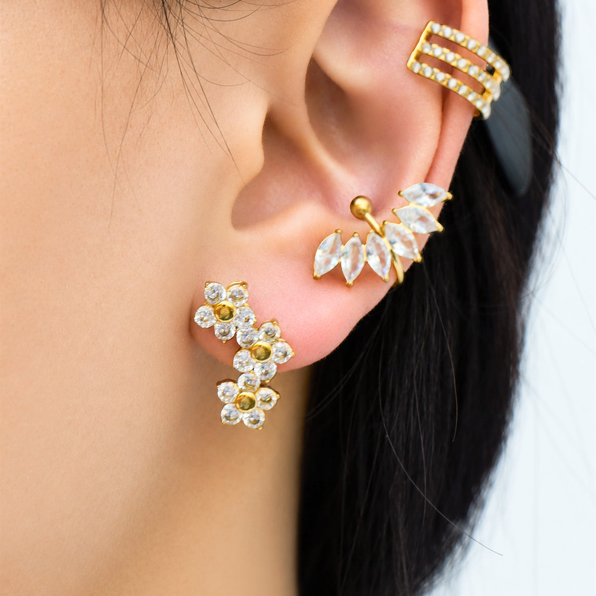 Model wearing gold three-flower cluster earrings with sparkling clear stones, styled alongside other gold ear jewelry