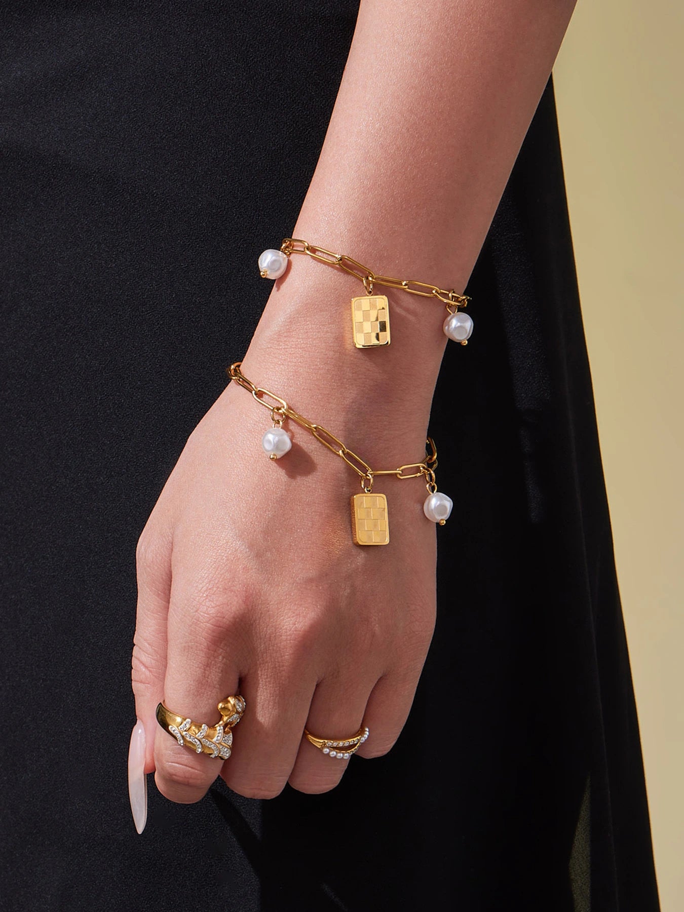 Two gold paperclip charm bracelets with pearl-look beads and checkerboard pendants on hand