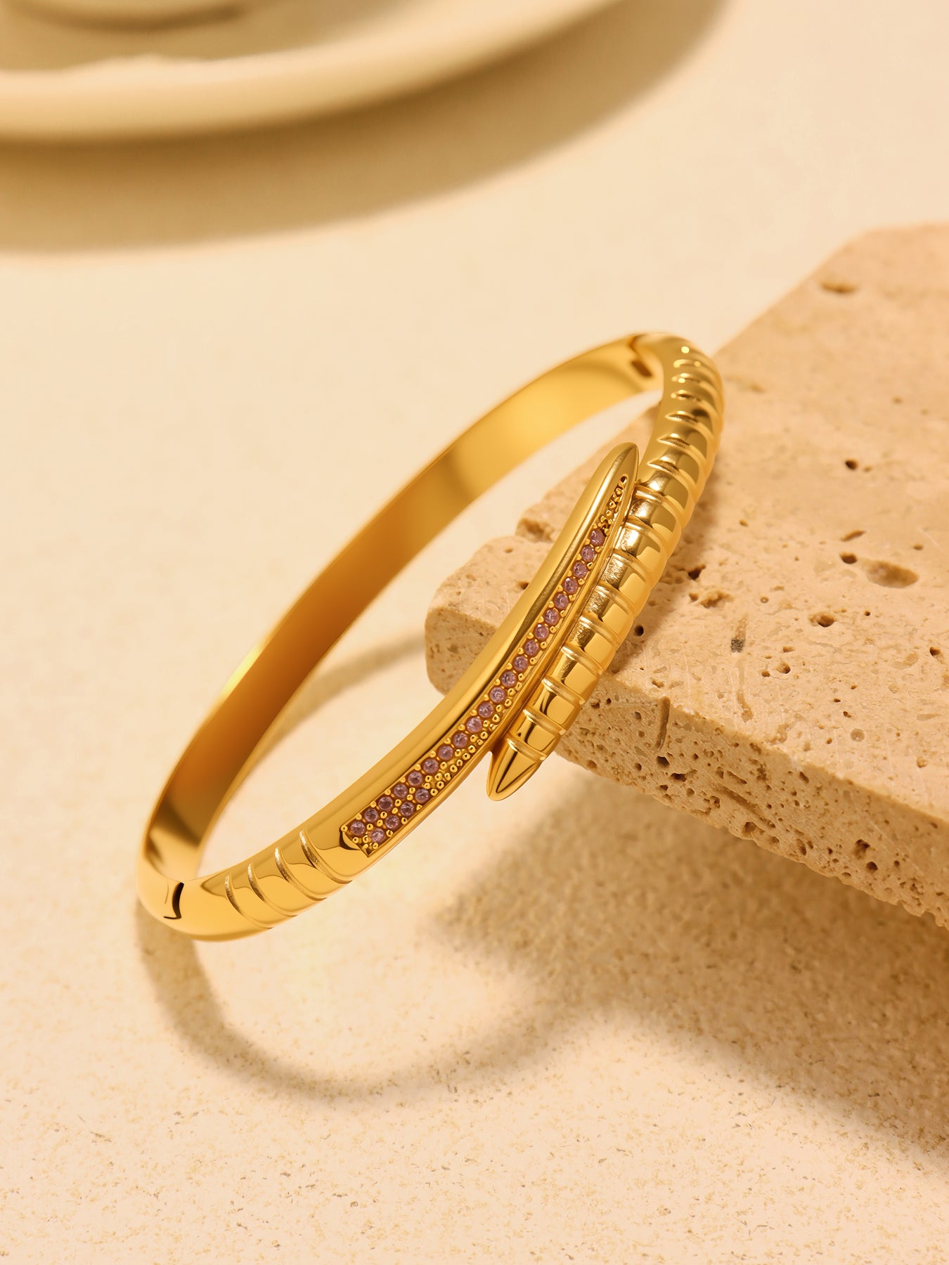 Gold-tone nail bangle bracelet with pink zircon accents resting against a beige stone block