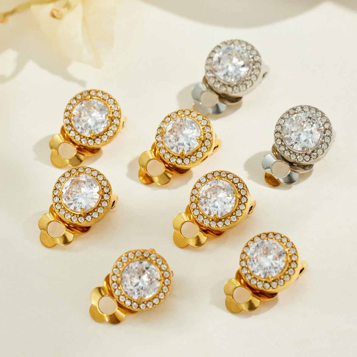 Assorted gold and silver round halo clip-on earrings arranged on a light surface, showing sparkling center stones and matching clip closures