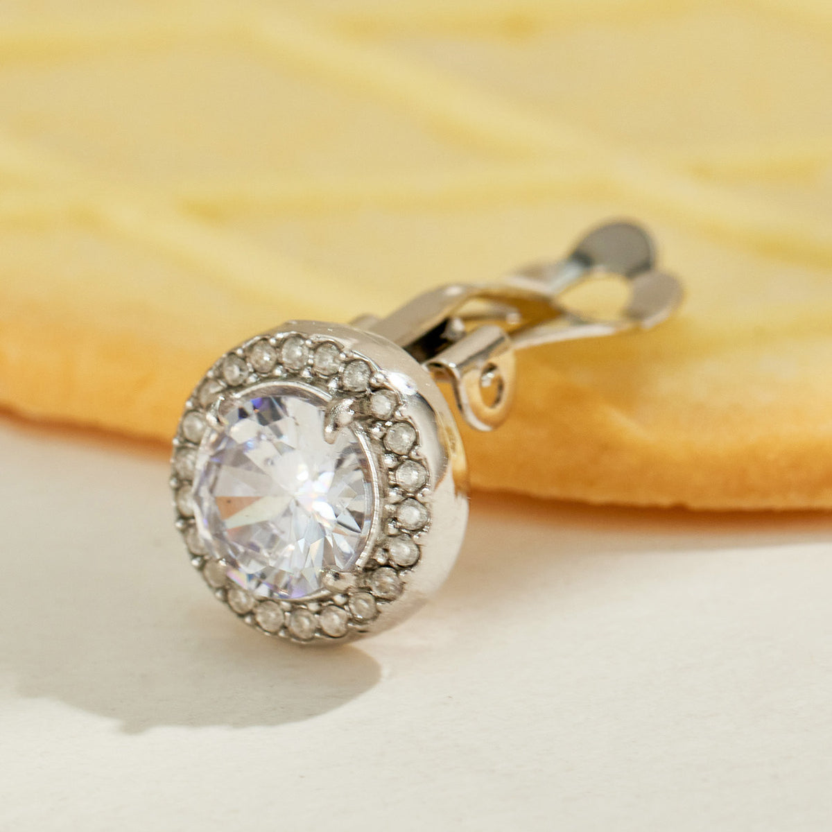 Silver-tone halo clip-on earring displayed against a soft yellow background, featuring a faceted clear center stone and pavé-style halo