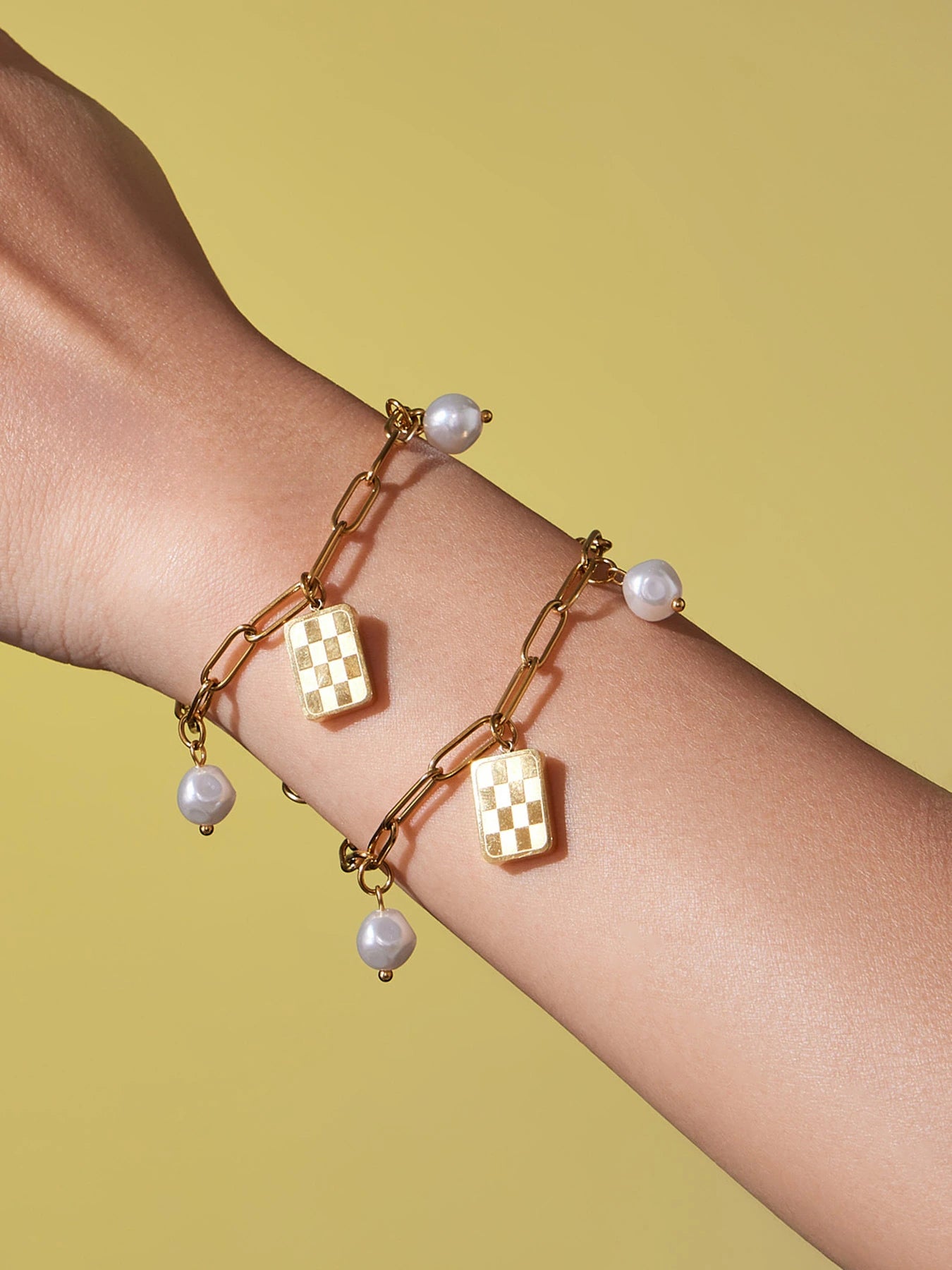 Close-up of matching gold paperclip charm bracelets with checkerboard and pearl-look charms