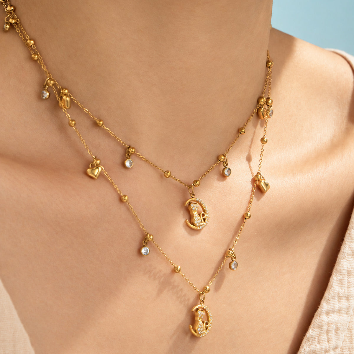 Two gold-tone stainless steel cat moon charm necklaces worn at different lengths, showing heart charms and clear stone accents