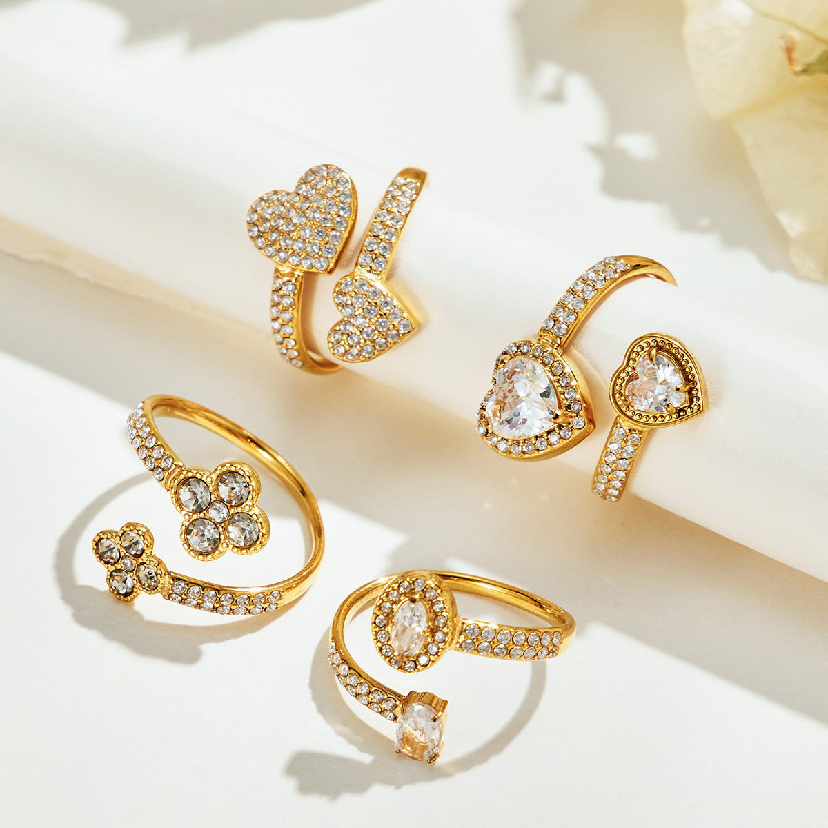 Four gold-tone stainless steel flower, heart and two-stone open rings displayed on a cream surface