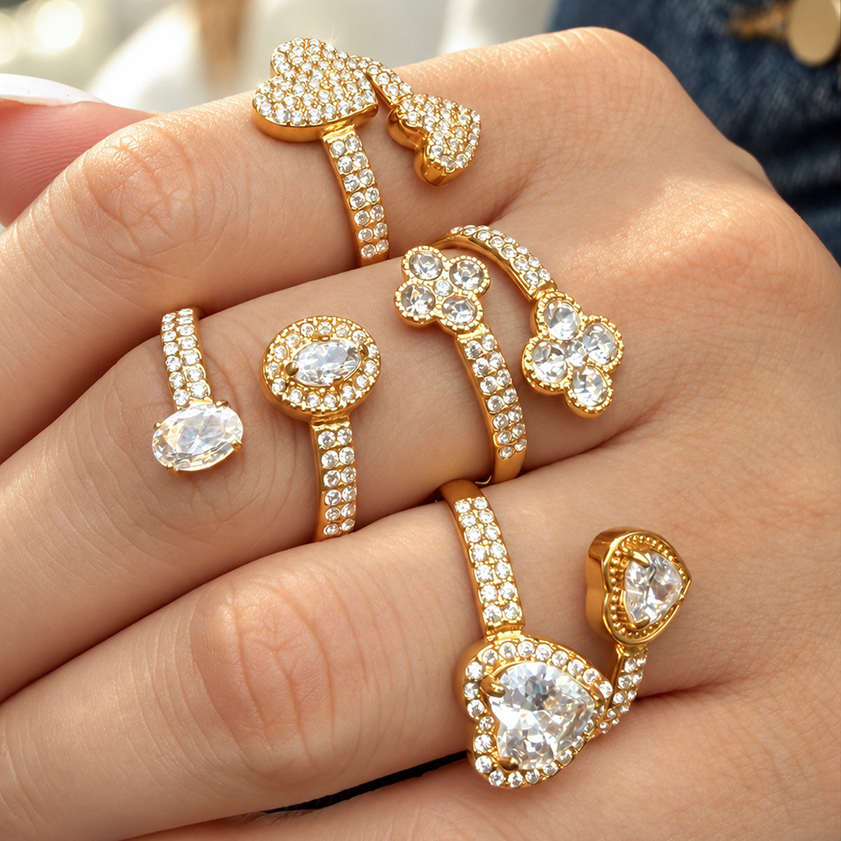 Four gold-tone stainless steel open ring designs worn across fingers, showing flowers, hearts and clear stones