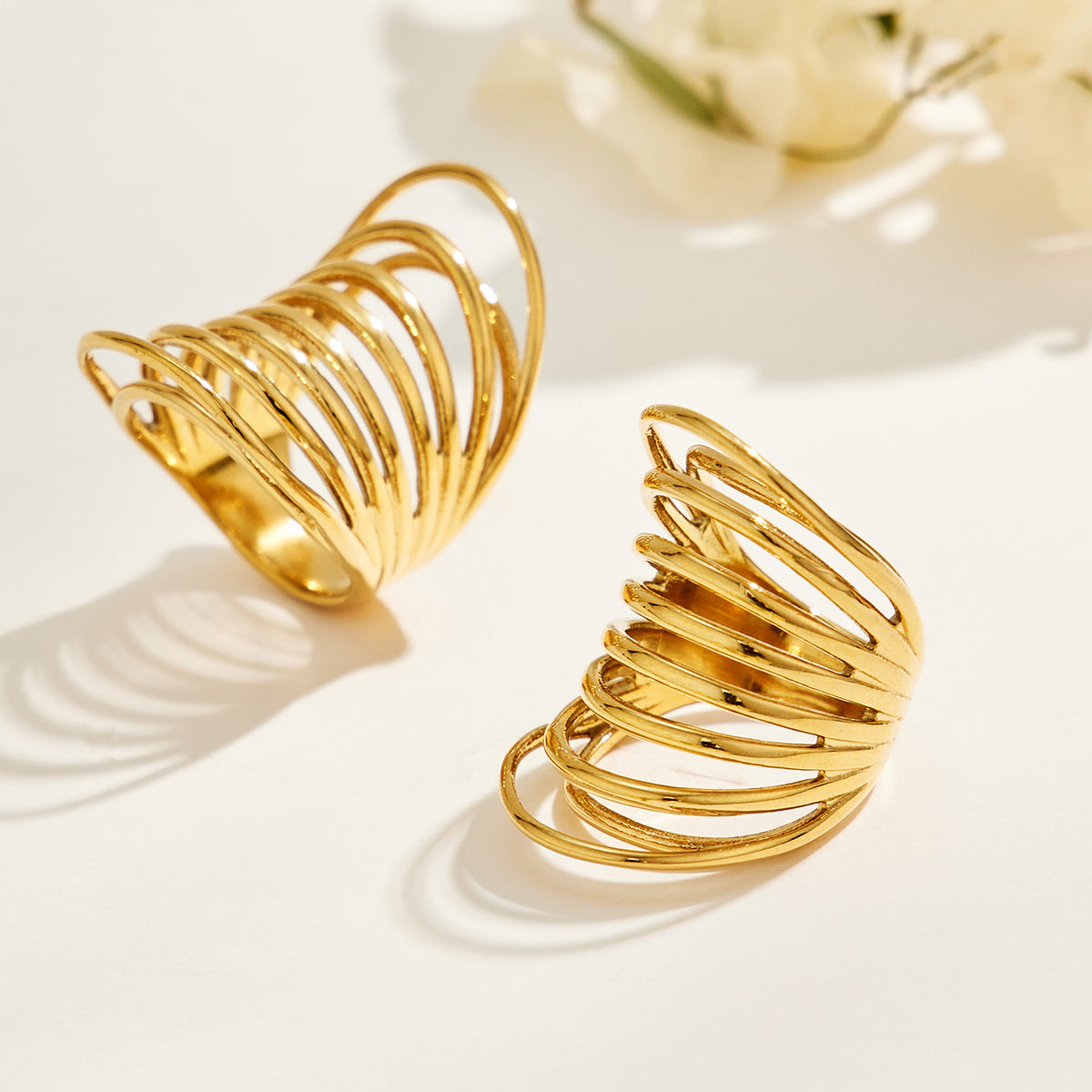 Two gold-tone stainless steel sculptural rings on a cream surface, casting shadows through their openwork bands