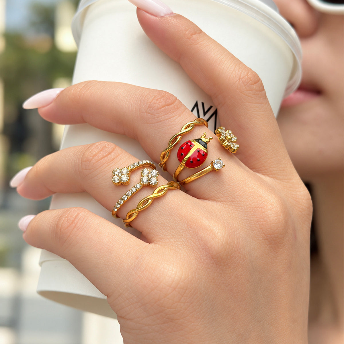 Gold-tone stainless steel ladybug and flower rings styled with twisted bands on a hand holding a white cup