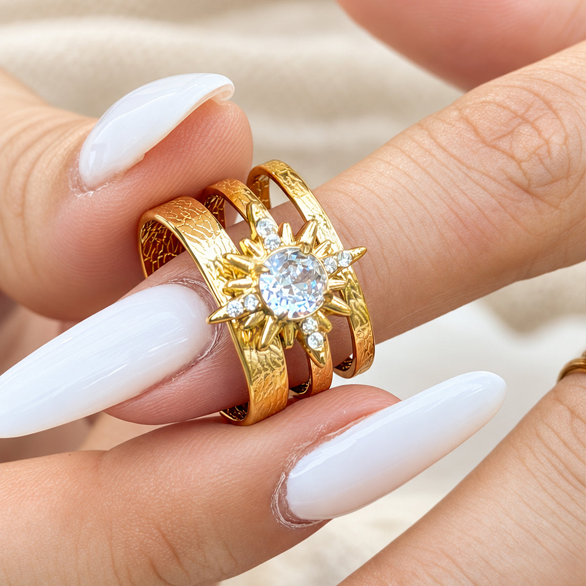 Close-up of a gold-tone stainless steel sunburst flower ring held between fingers with white nails