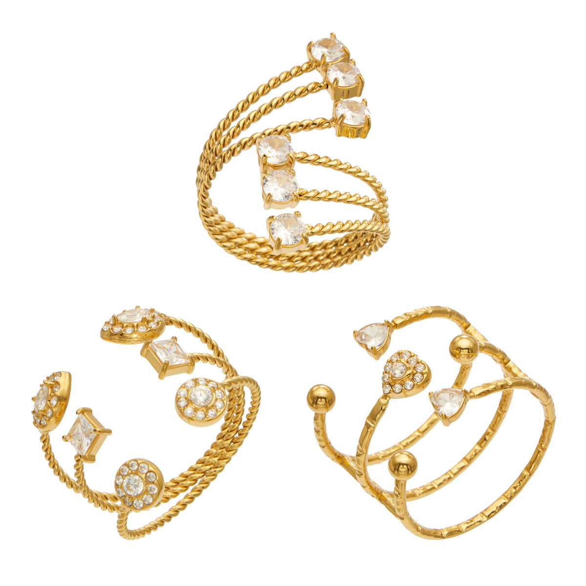Three gold-tone stainless steel triple-band open rings with heart, geometric and clear stone-tip designs on a white background