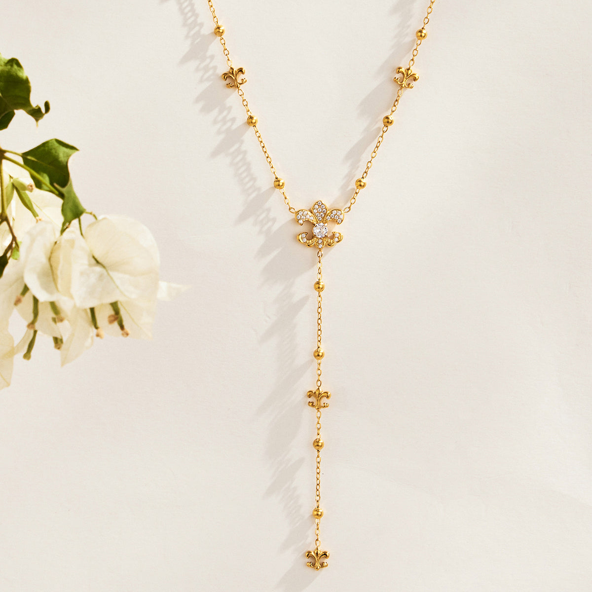 Gold-tone stainless steel Y lariat necklace with ornamental motifs and a beaded chain drop beside white flowers