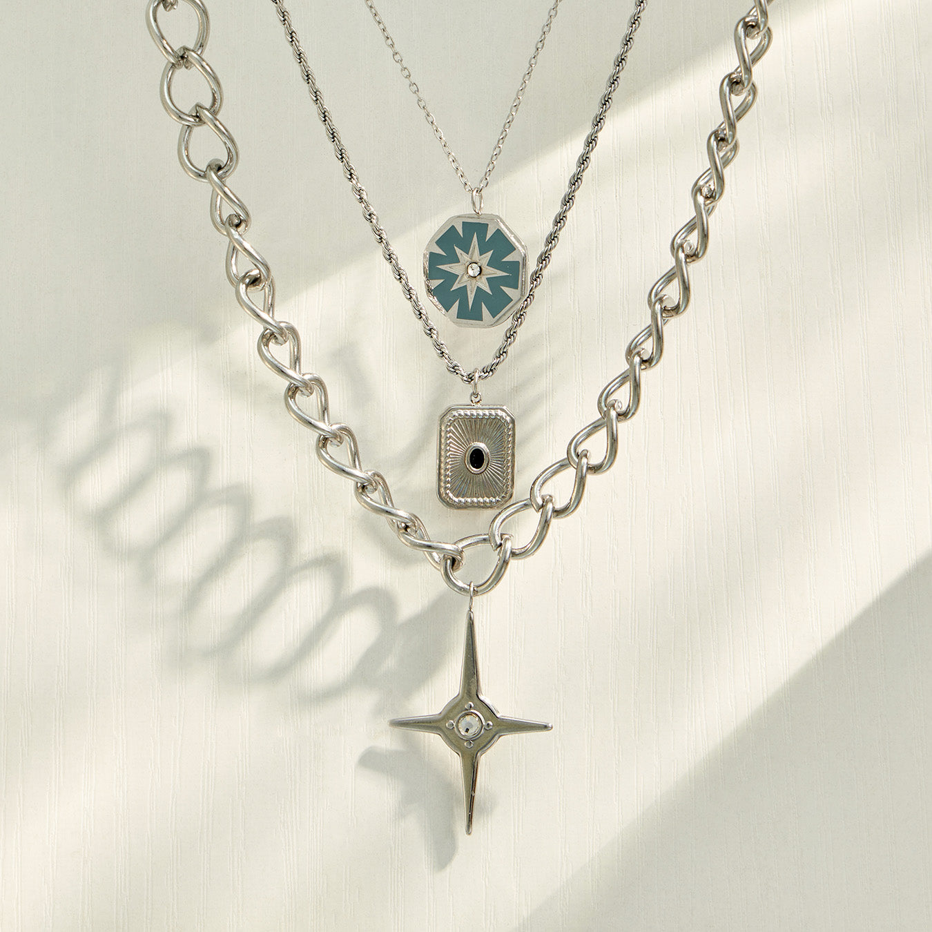 Three steel-tone geometric pendant necklaces arranged in a layered flat-lay display