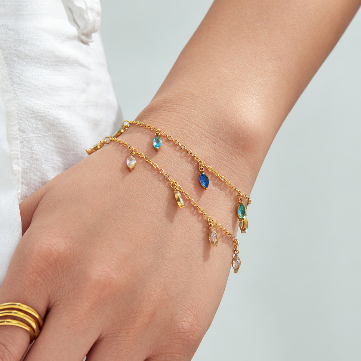 Gold-tone chain bracelets with colorful dangling stone charms styled on a hand with gold rings.