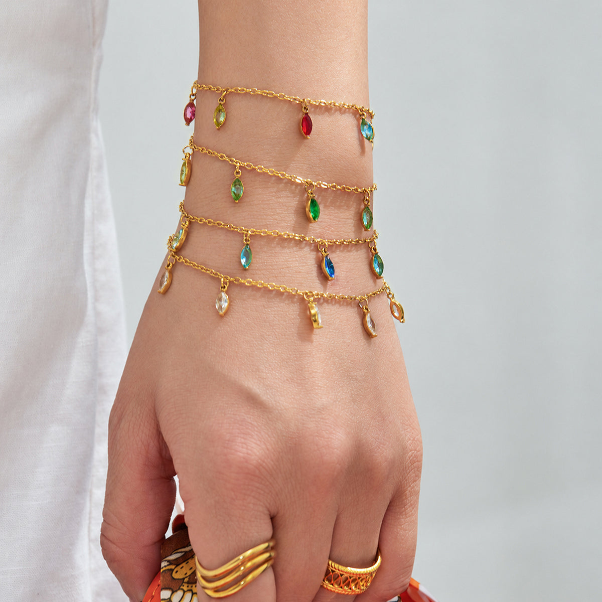 Multiple delicate gold-tone teardrop charm bracelets layered on a model's wrist against white clothing.