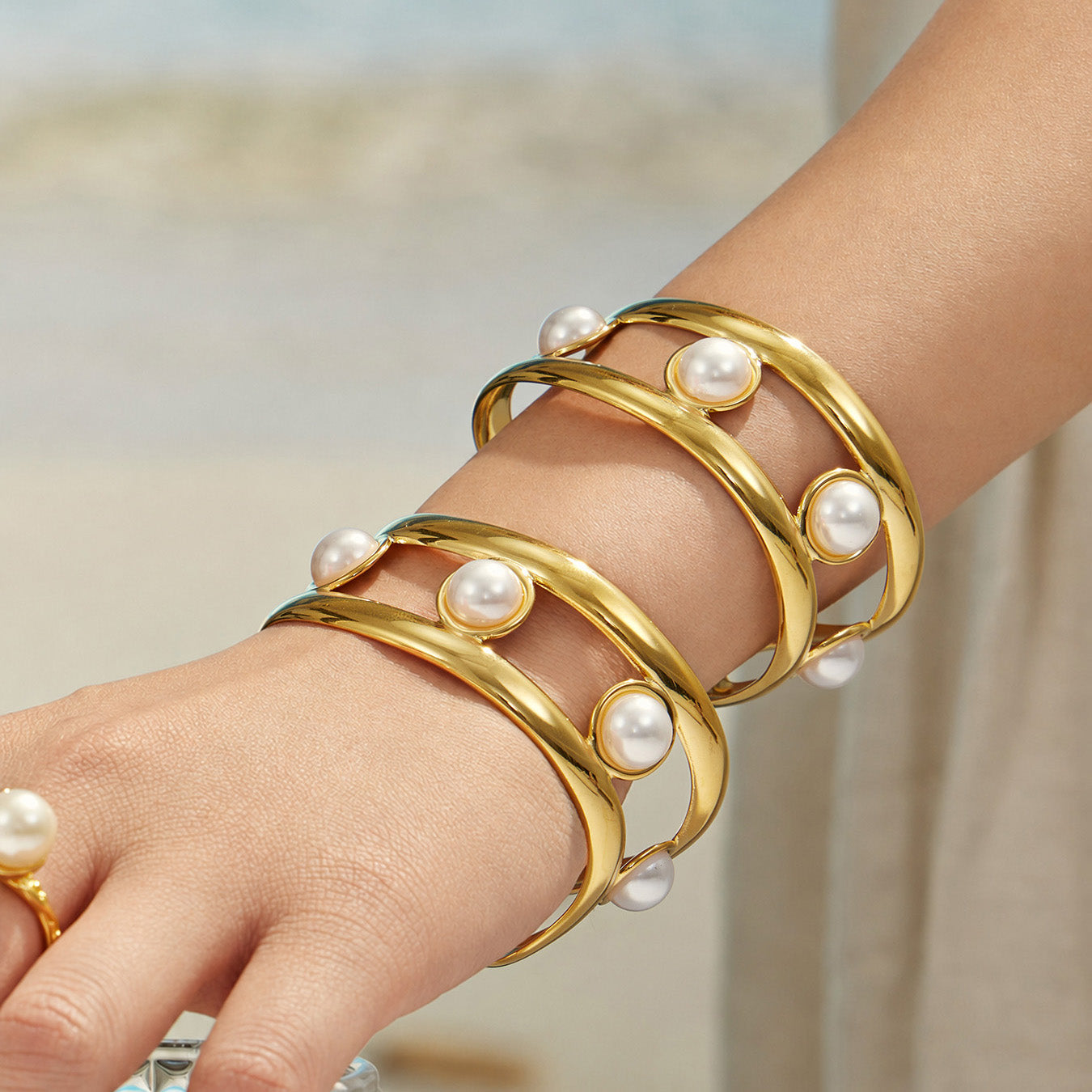 Wide gold pearl cuff bracelet worn on a model wrist near the beach
