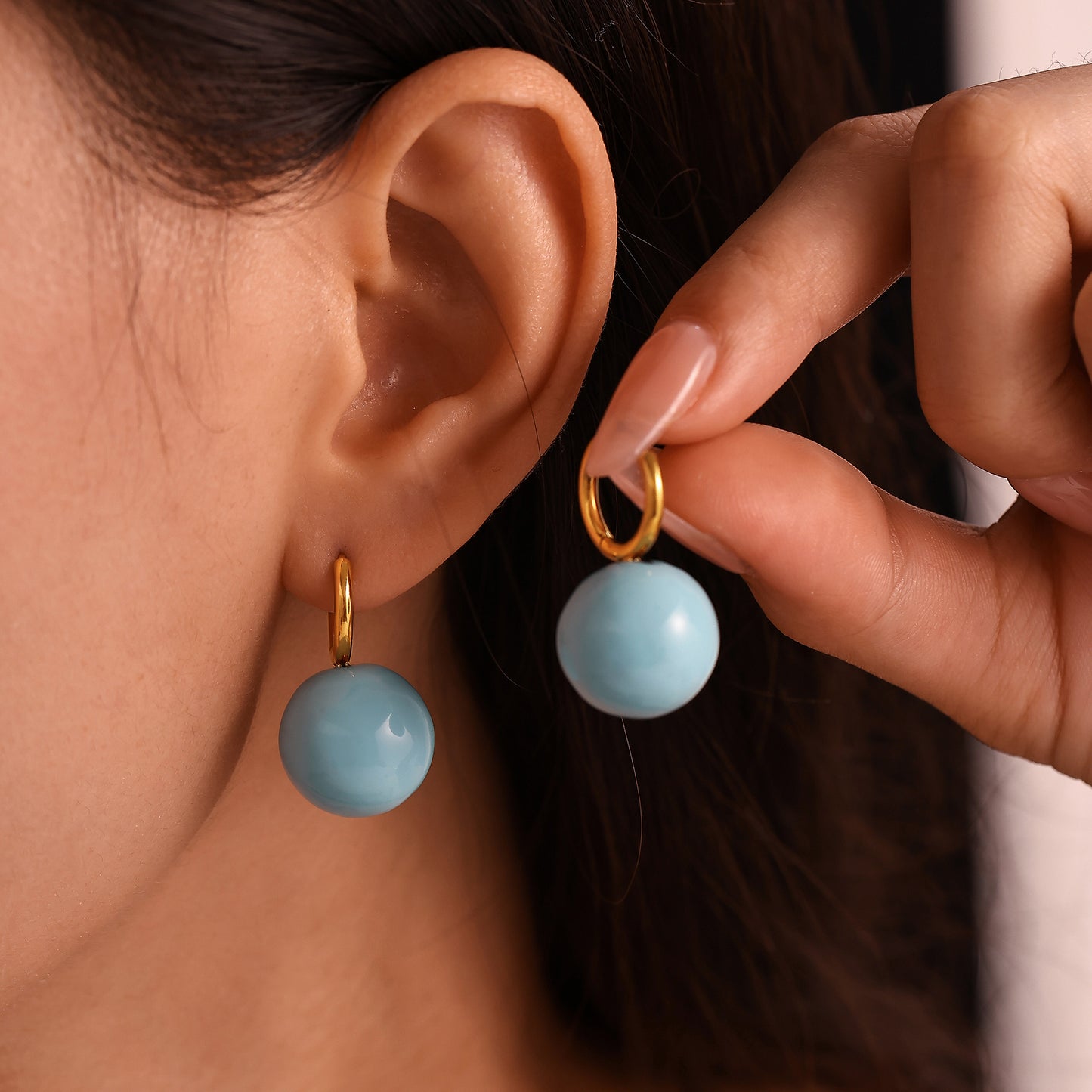 Model wearing and holding light blue ball drop earrings with gold-tone hoops