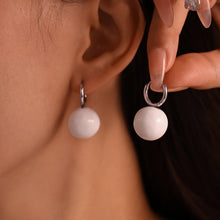 Model wearing and holding white ball drop earrings with steel-tone hoops