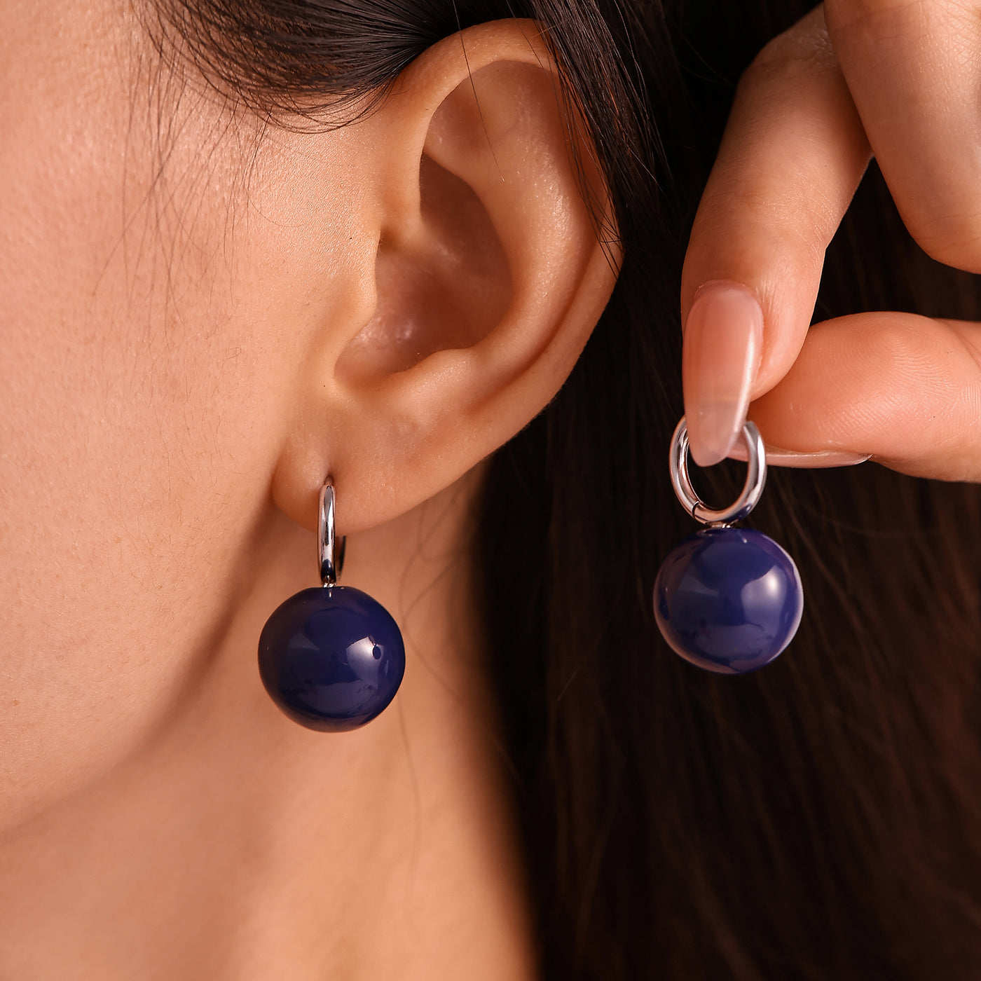 Model wearing and holding navy ball drop earrings with steel-tone hoops