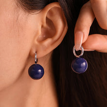 Model wearing and holding navy ball drop earrings with steel-tone hoops