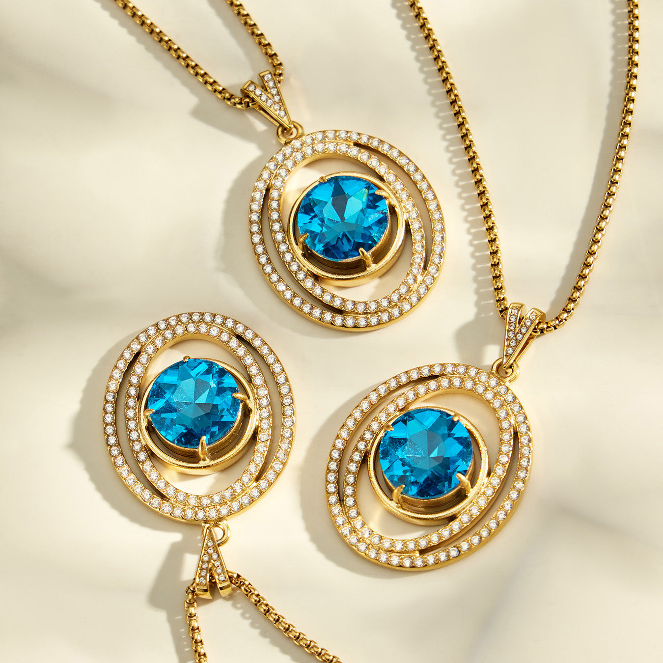 Three blue glass halo pendants with gold-tone chains arranged on a cream surface
