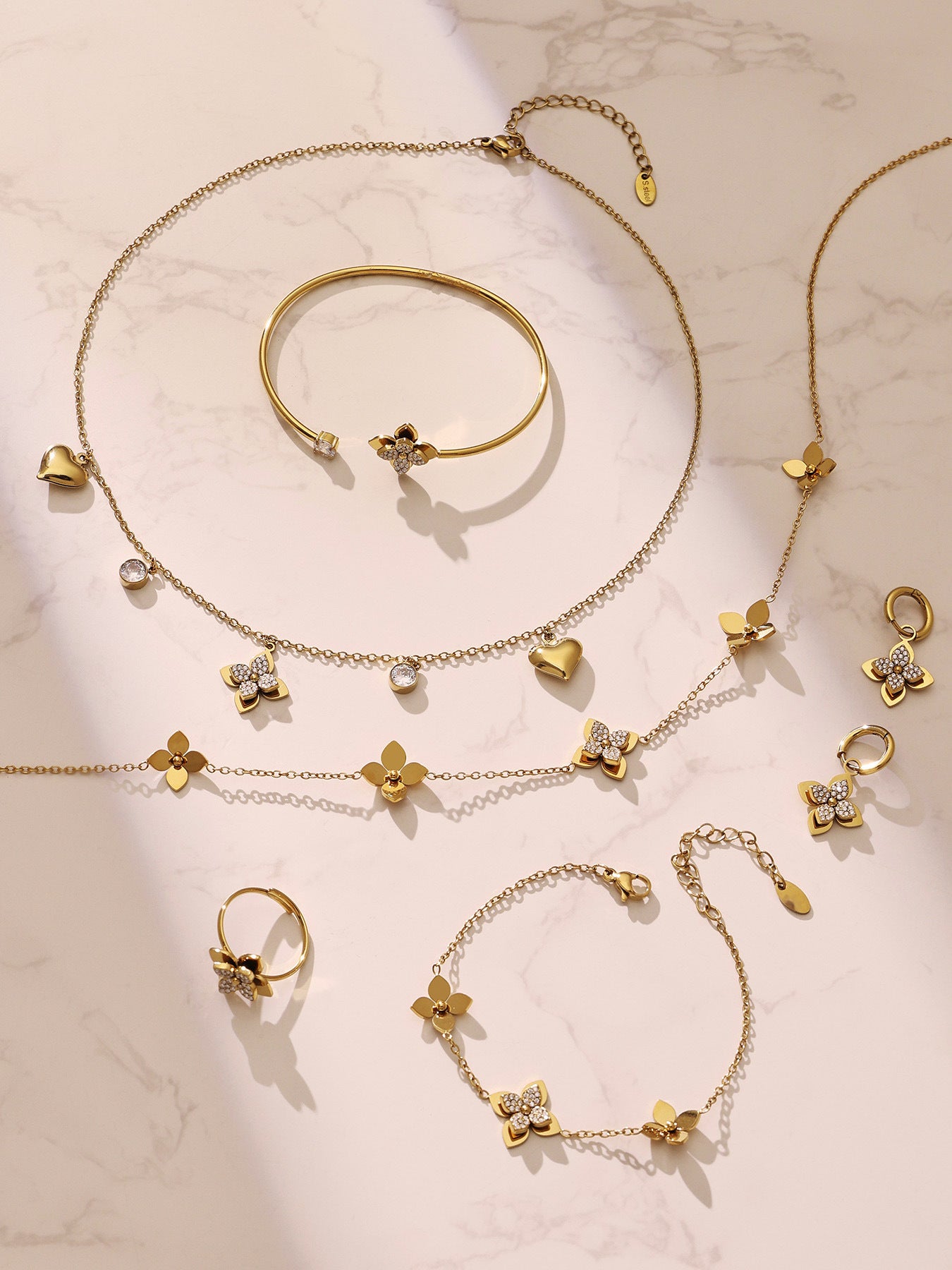 Aesthetic flat lay of the four-petal flower jewelry collection including matching bracelets and necklaces in a rich gold tone.