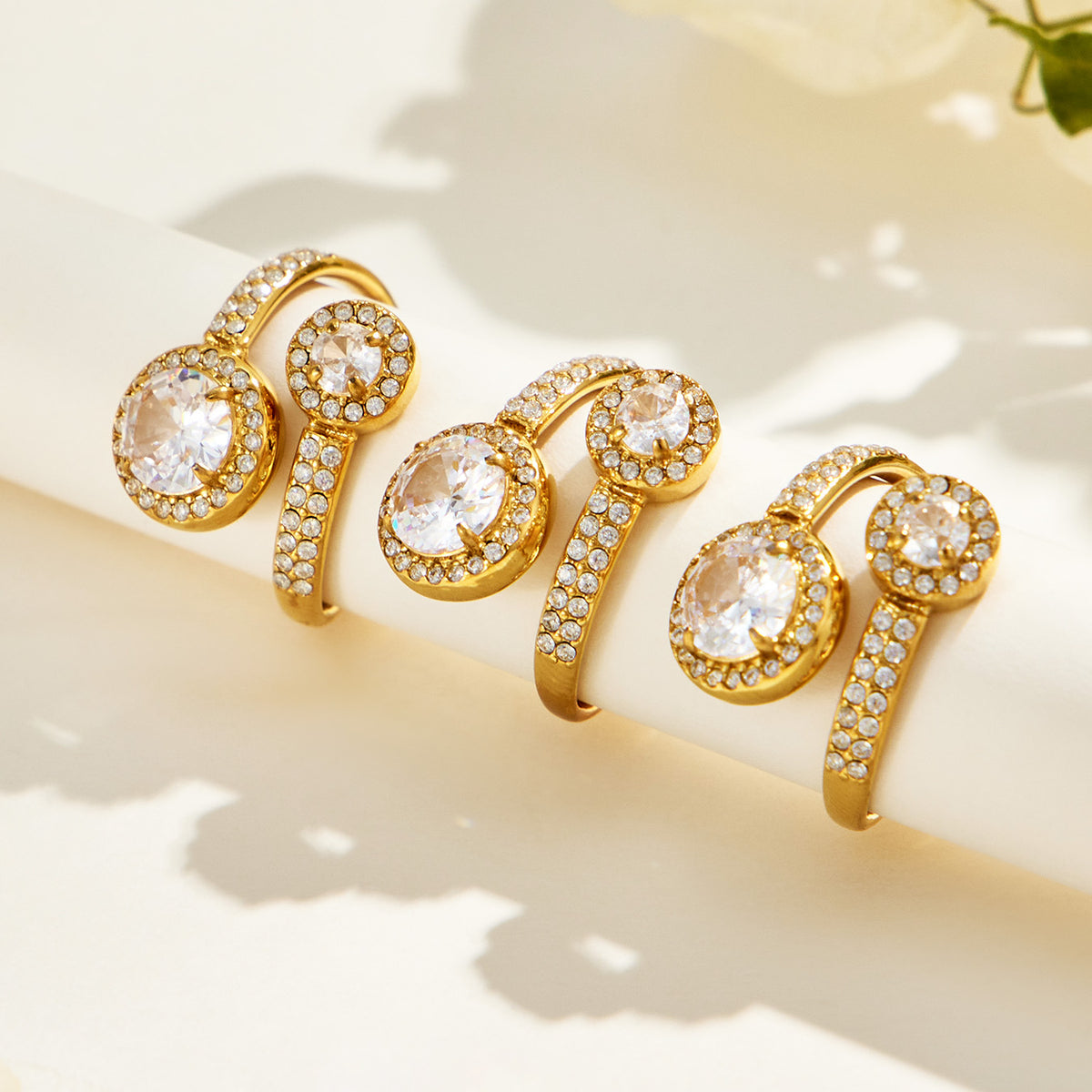 Three gold-tone stainless steel double halo rings arranged on a cream surface with the round stone settings facing forward
