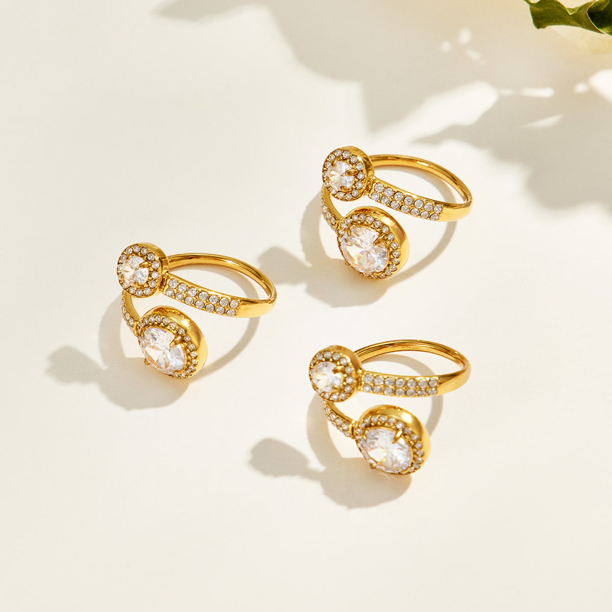 Three gold-tone stainless steel double halo rings displayed at different angles, showing the round stone settings and open bands