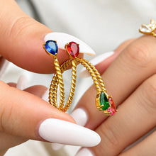 Gold-tone stainless steel twisted open rings held between fingers, showing blue, red, pink and green teardrop stones