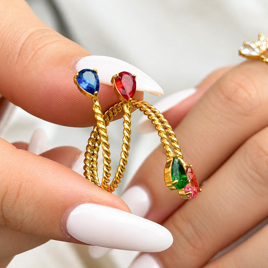 Gold-tone stainless steel twisted open rings held between fingers, showing blue, red, pink and green teardrop stones