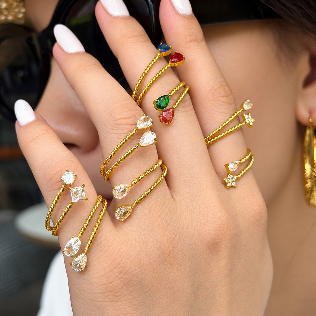 Model wearing several gold-tone stainless steel twisted open rings with star, square and teardrop stone details