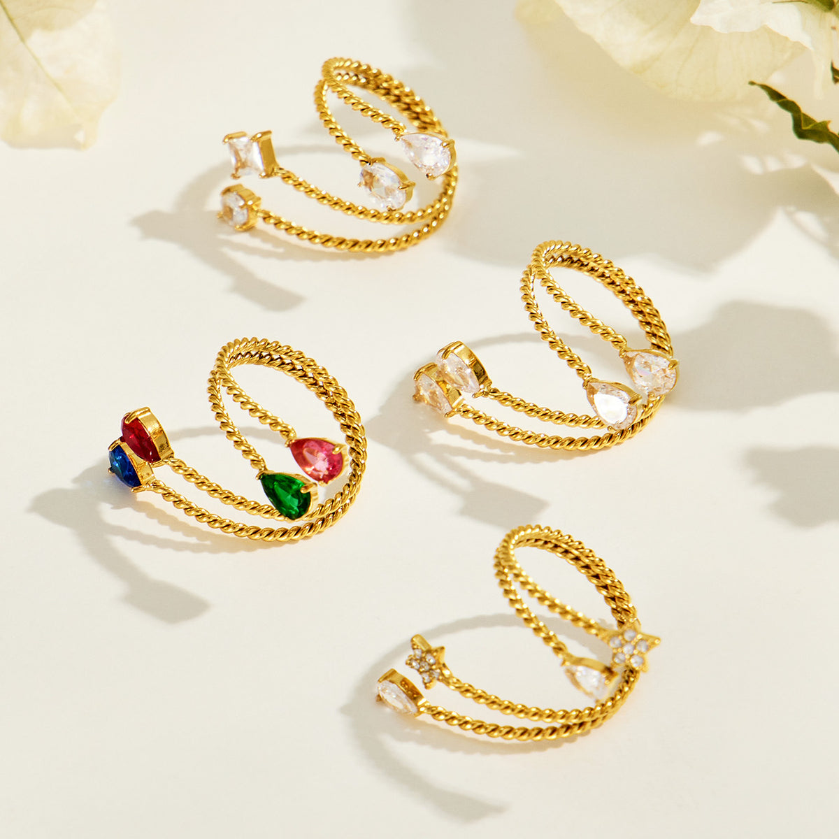 Gold-tone stainless steel twisted open rings with clear and multicolor stones displayed among pale petals
