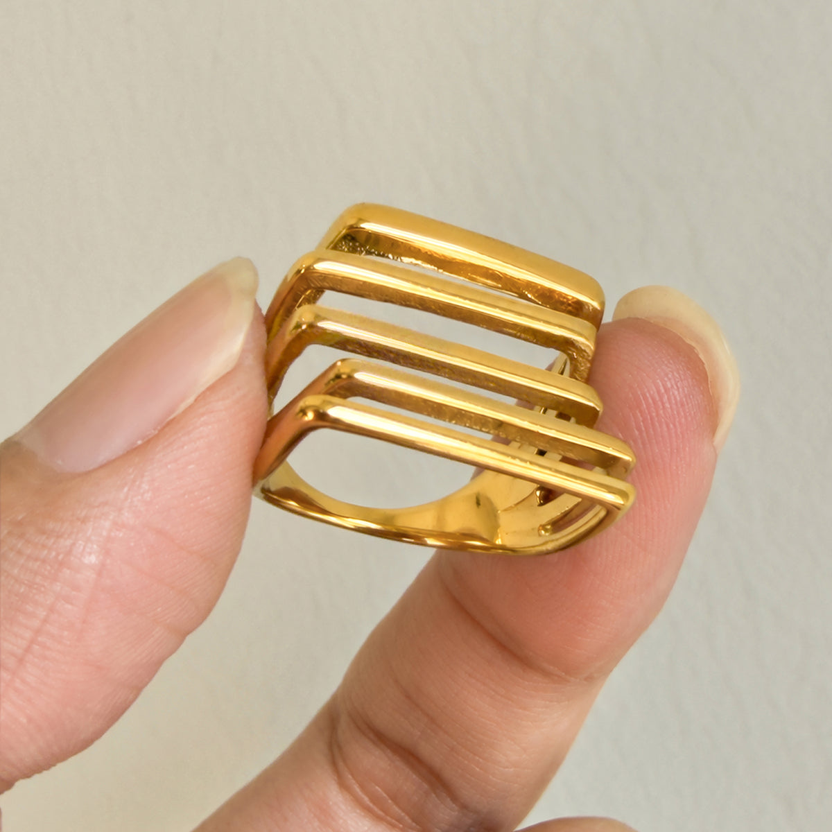 Gold-tone stainless steel openwork ring held between two fingers, showing the curved band and multi-row face