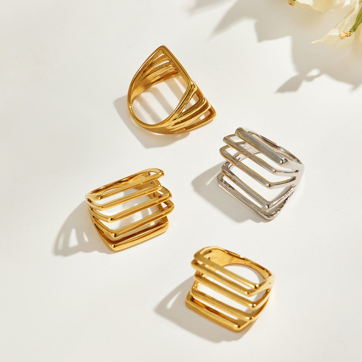 Three gold-tone rings and one steel-tone stainless steel multi-row ring arranged on a light background with floral shadows