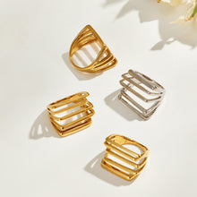 Three gold-tone rings and one steel-tone stainless steel multi-row ring arranged on a light background with floral shadows