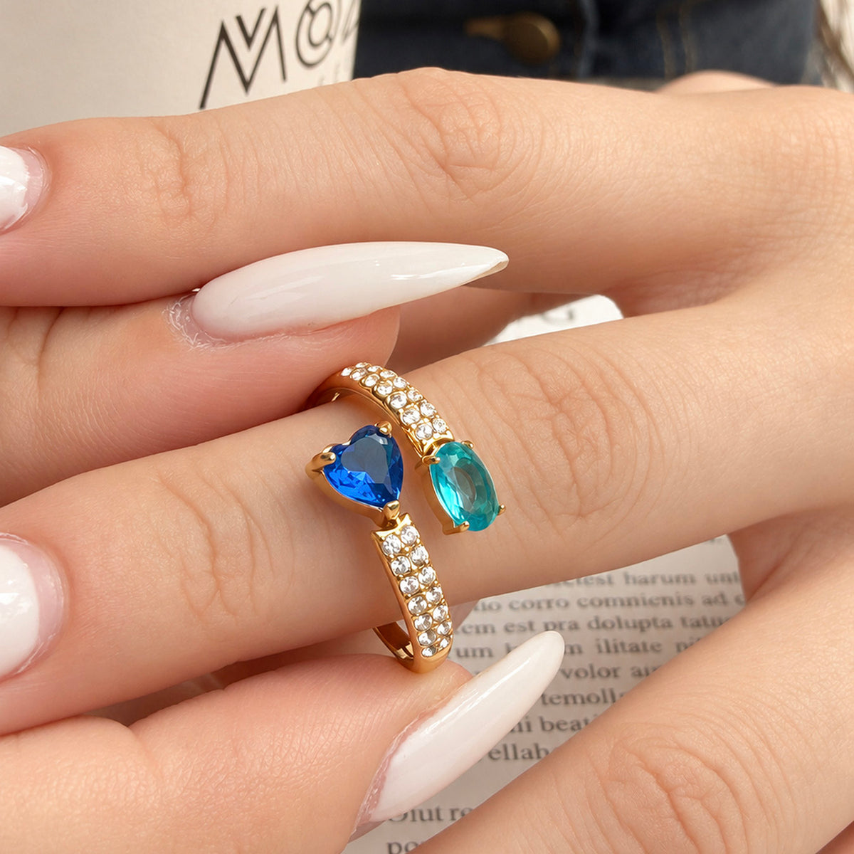 Gold-tone stainless steel open ring worn on a finger with a blue heart stone, teal oval stone and clear-stone band