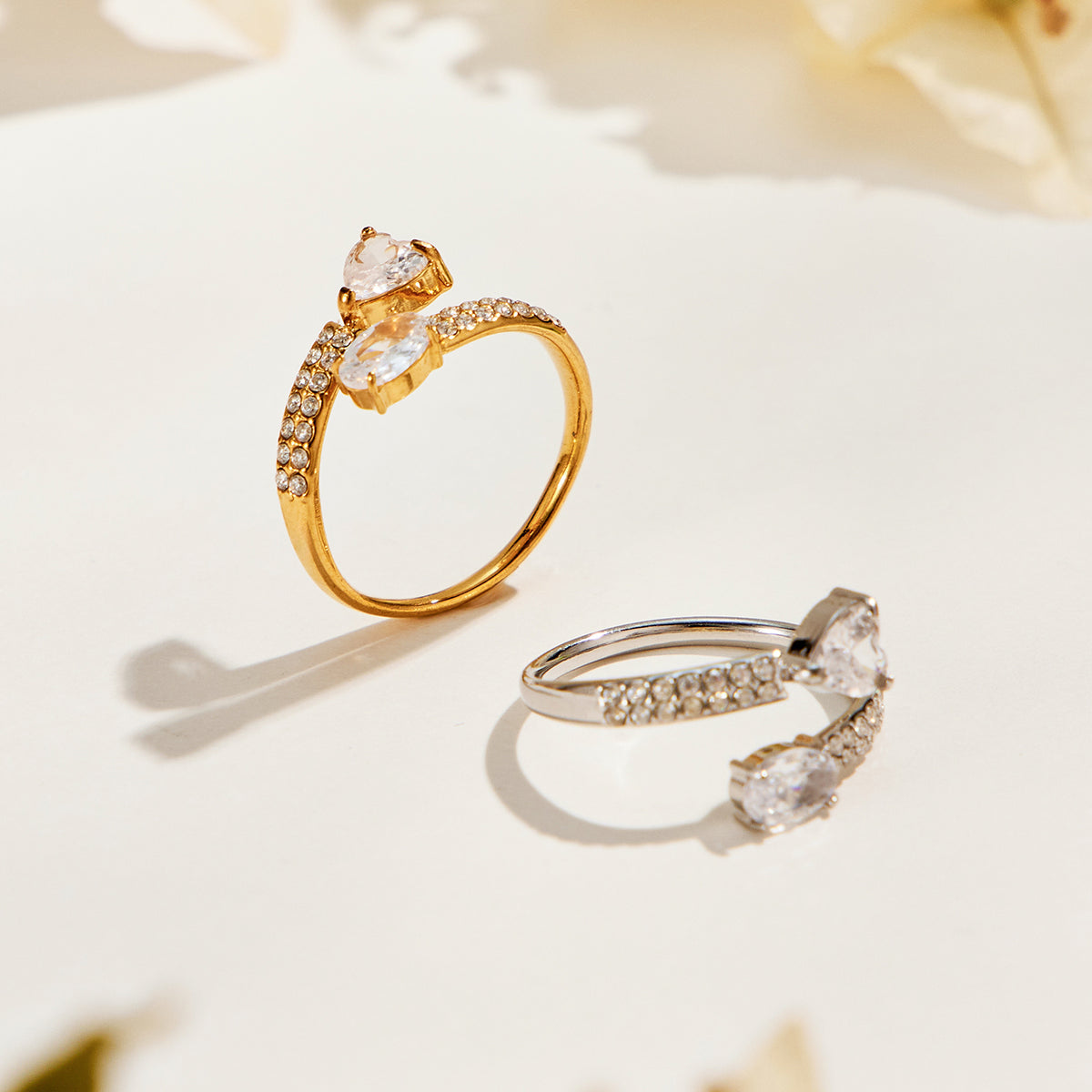 Gold-tone and steel-tone stainless steel heart and oval rings with clear stones displayed together on a cream surface