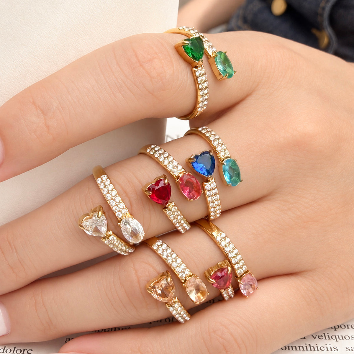 Front view of gold-tone stainless steel heart and oval rings on fingers, with green, blue, red, pink, clear and champagne stones