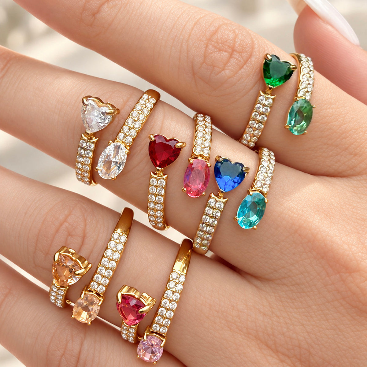 Close-up of gold-tone stainless steel rings worn on fingers, highlighting the heart and oval stones and rows of small clear stones