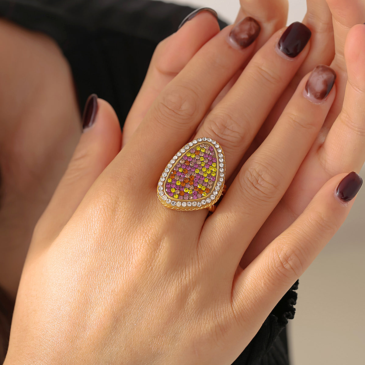 Top view of a hand with burgundy nails wearing a gold-tone teardrop ring with colorful stones and a clear stone border.