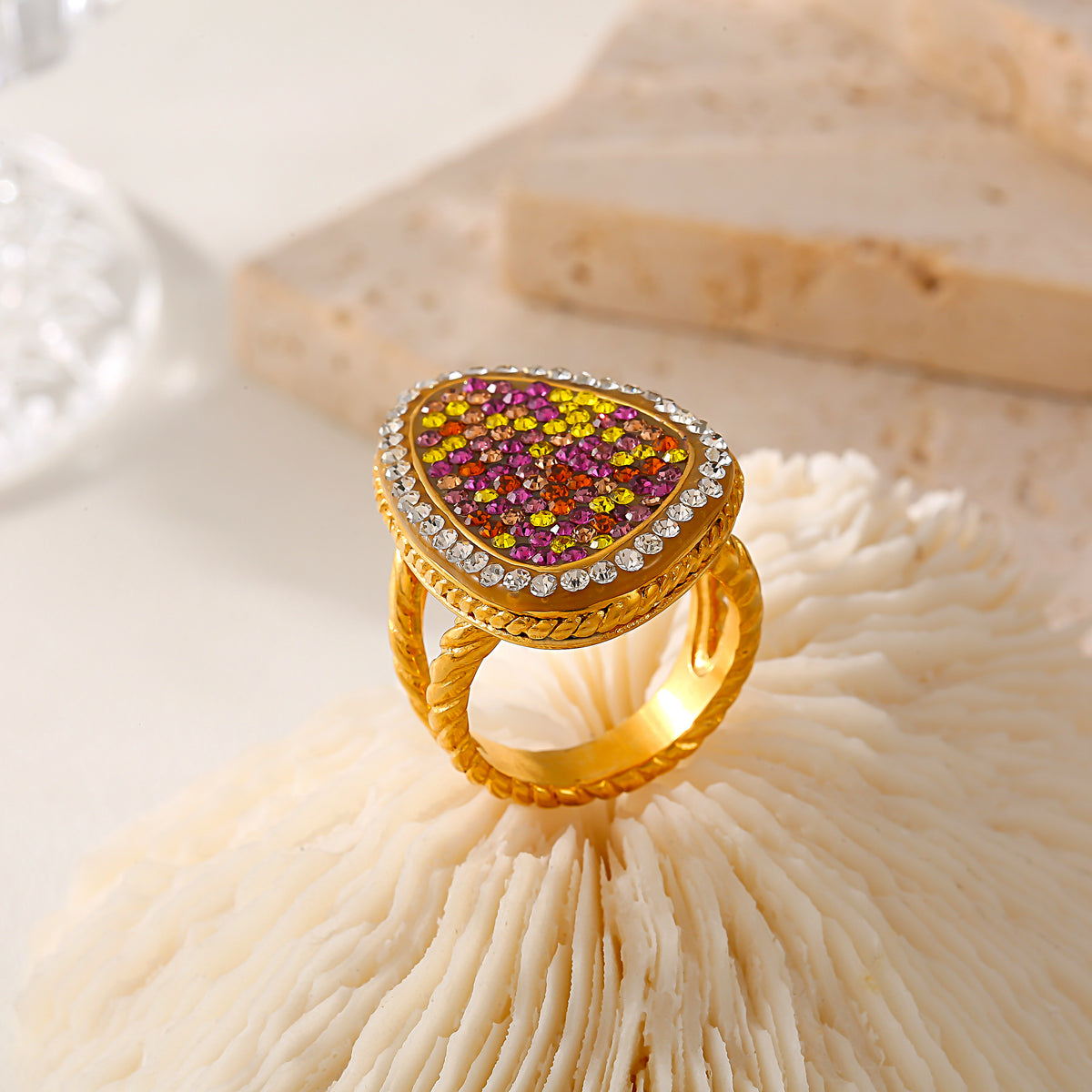 Gold-tone A907 teardrop ring standing on a cream textured display, showing the colorful stone face and curved band.
