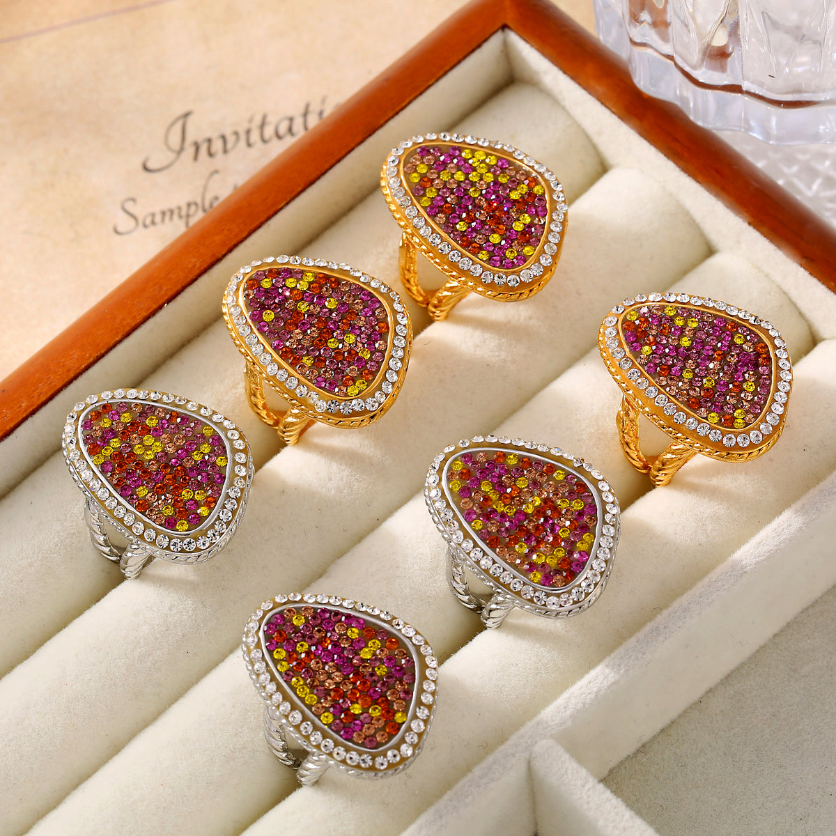 Six multicolor teardrop statement rings in gold-tone and steel-tone settings arranged in a cream jewelry display case.