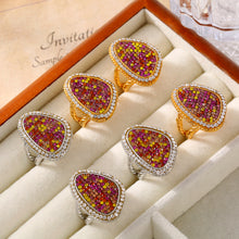 Six multicolor teardrop statement rings in gold-tone and steel-tone settings arranged in a cream jewelry display case.